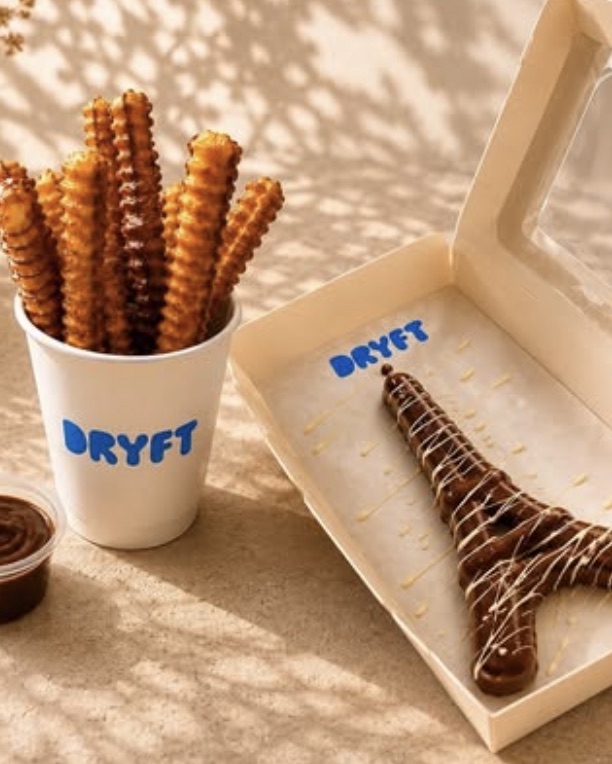 A cup of waffle fries next to an Eiffel Tower waffle in a Dryft box, covered in chocolate and white chocolate drizzle.